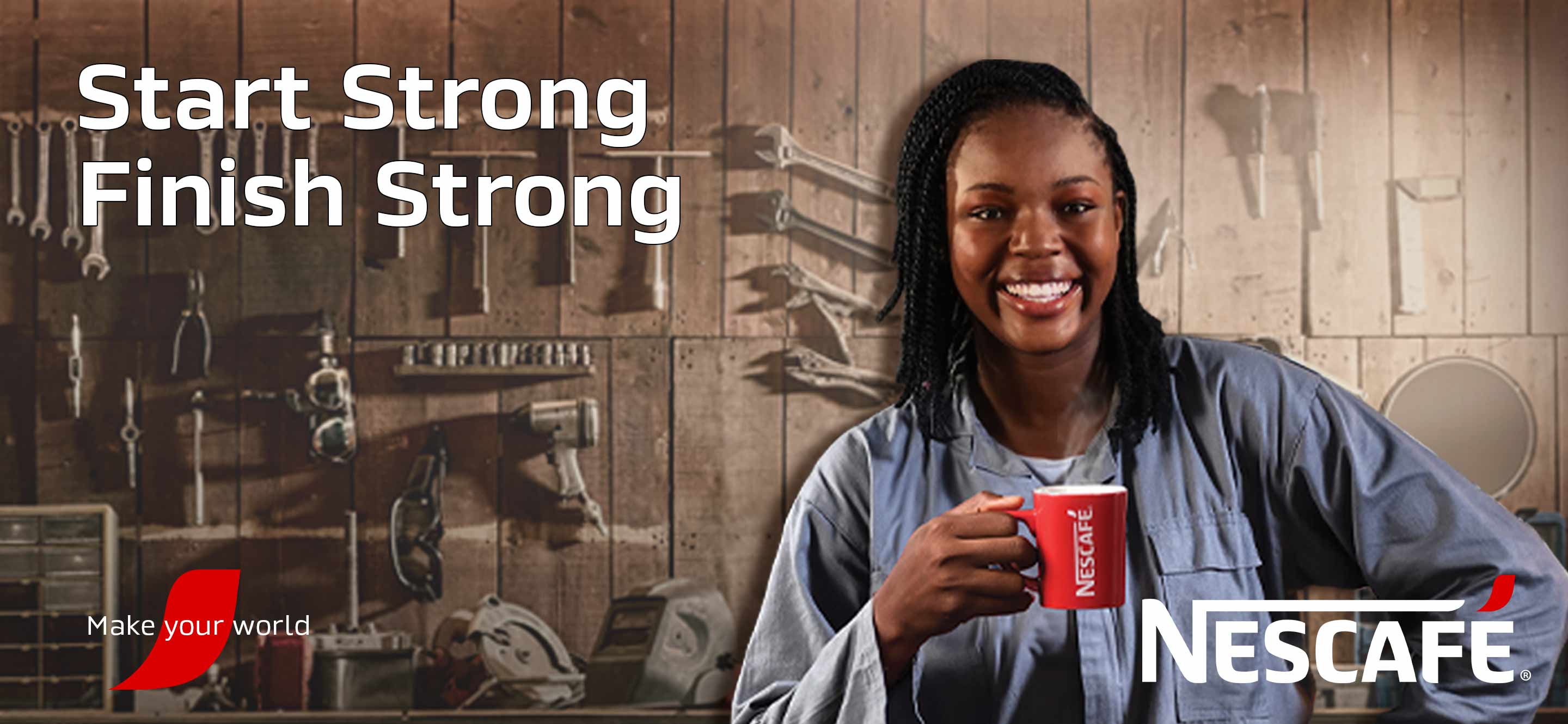 A woman in a blue jumpsuit with a big smile is standing in front of a wooden wall with several workshop tools while holding a red NESCAFÉ mug.