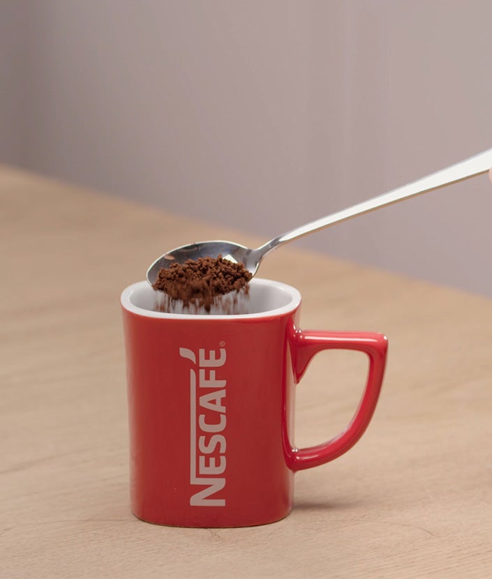 Dodavanje kašičice Nescafé Black Roast kafe u crvenu Nescafé šoljicu