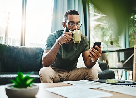 A man drinking coffee looking at his phone while on a sofa