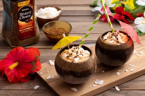Dos cocos con café cremoso, crema batida, coco rallado, pajillas y flores tropicales