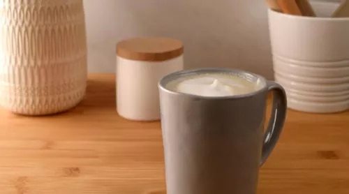 Photograph: gray ceramic mug with frothy coffee on wooden table, neutral jars in background