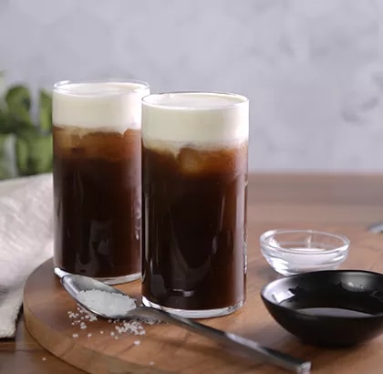 Two tall iced coffees with creamy foam on a wooden board, spoon and small bowls nearby