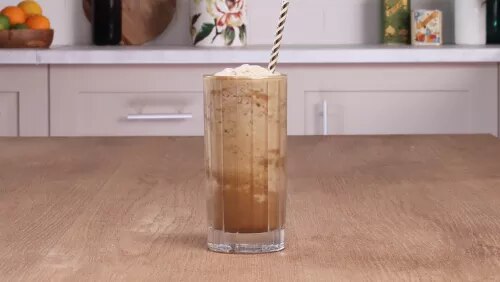 Vaso alto de café helado con espuma y pajilla sobre mesa de madera, fondo de cocina desenfocado
