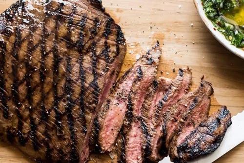 Photo: sliced grilled steak, medium-rare, charred, on wooden board with green sauce