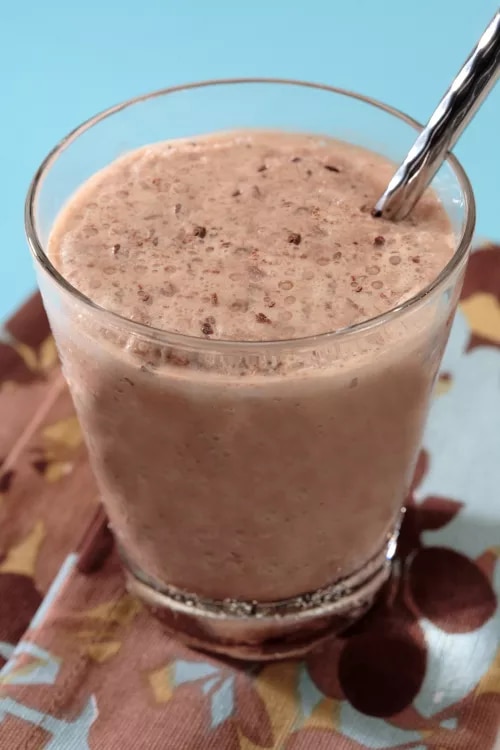 Photograph of a glass of chocolate-speckled smoothie with spoon on patterned napkin