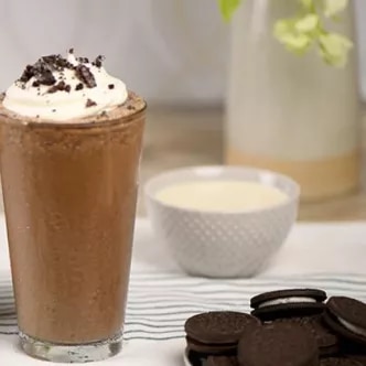 Fotografía de batido de chocolate con crema y trozos de galleta; tazón y galletas al lado.