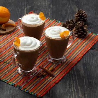 Fotografía: tres vasos de chocolate caliente con crema y cáscara de naranja, mantel rayado y piñas