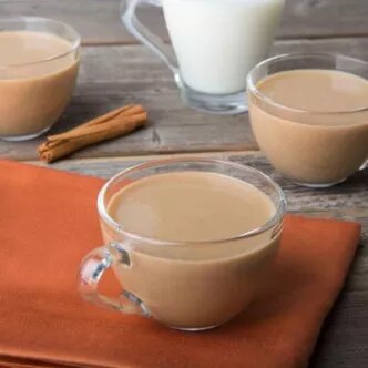Fotografía: tazas de vidrio con té con leche, palitos de canela y jarra sobre mesa rústica