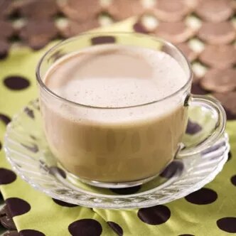 Foto: taza de vidrio con bebida cremosa sobre platillo, sobre mantel verde con lunares marrones