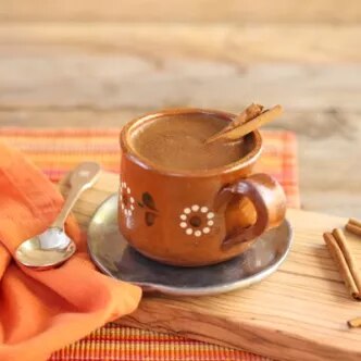 Fotografía de taza de café espumoso con una rama de canela, sobre platillo y servilleta naranja.